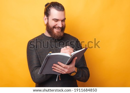 Handsome cheerful young man writing in agenda or planner over yellow background.