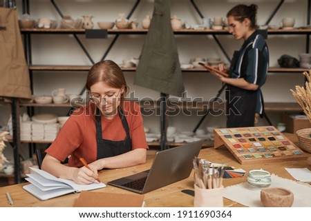 Portrait of female small business owner taking notes in planner while working at wooden table with laptop in pottery workshop, copy space
