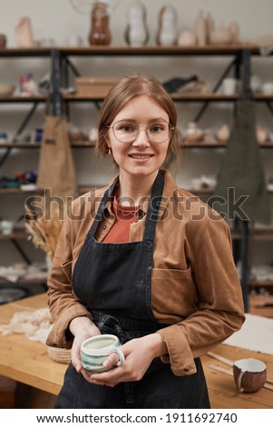 Vertical waist up portrait of young female potter looking at camera while posing in workshop, hobby and small business