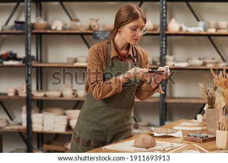 Portrait of young woman taking smartphone photo of raw ceramic bowl in pottery workshop to post on social media, small business concept, copy space