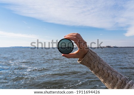 The arm of a woman holding up a lens filter in front of the Statue of Liberty