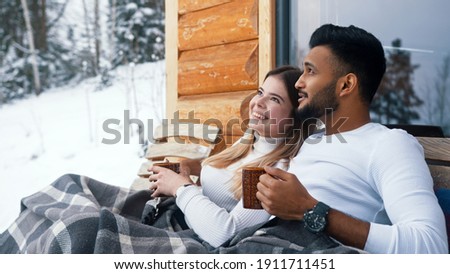  Winter holidays. Multiracial young couple sitting on the bench and kissing outdoors. Drinking hot beverage. High quality photo