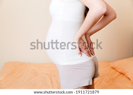 beautiful pregnant woman touching her tummy and keeping one hand on her back at home on bed.