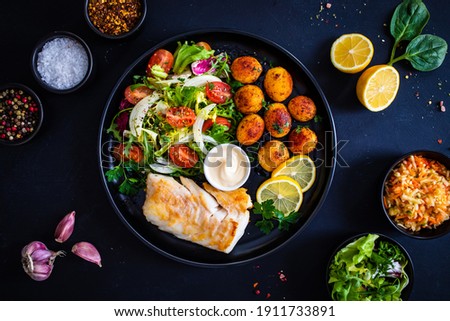 Fish dish - fried cod fillet with potatoes and vegetable salad on black wooden table 
