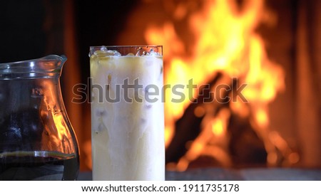 Milk is pouring into a Glass of coffee with ice in front of burning fireplace in a country house.