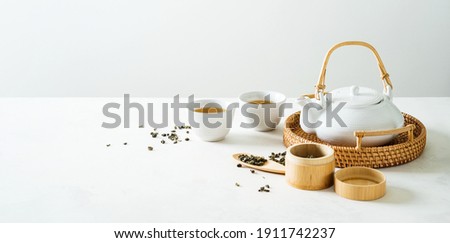 Asian tea concept mockup, two white cups of tea and teapot surrounded with green dry tea  with space for a text on light background. Wide banner. Royalty-Free Stock Photo #1911742237