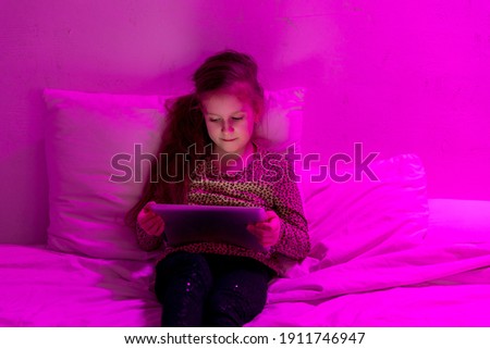 Little girl watching cartoon on tablet in bed in the evening. She looks closely at the screen and smiles. Home entertainment. Evening twilight. High quality photo