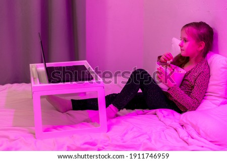 Little girl watching cartoon on laptop in bed in the evening and eating popcorn. Home entertainment. Evening twilight. High quality photo