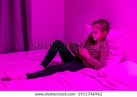 Little girl watching cartoon on tablet in bed in the evening. She looks closely at the screen. Home entertainment. Evening twilight. High quality photo