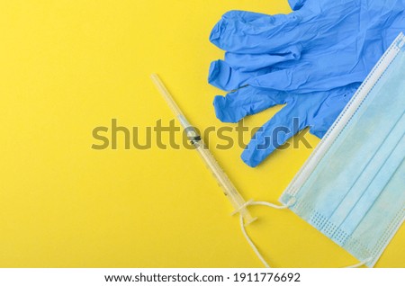 Medical glove with syringe and medical face mask on yellow background. Top view and selective focus.