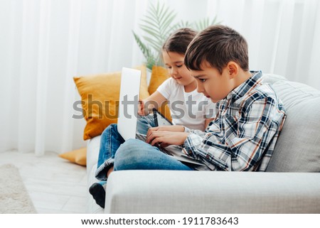 happy children sitting with crossed legs on a gray sofa in a nursery and looking at a laptop monitor   
