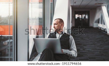 A portrait of a serious relaxed elegant man entrepreneur with a laptop, sprawled on a bench of a modern office hall while looking outside the window on a cityscape; a copy space place on the right