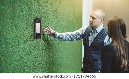 A man entrepreneur is showing and explaining to his female colleague how to use a terminal of a smart office system to change climate and illumination settings using a touch screen on a green wall