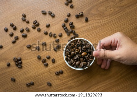 Selective focus of hand holding a cup full with coffee beans on wooden background.