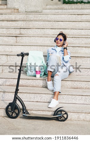 Beautiful young woman sitting on steps next to her electric scooter, listening to podcast in headphones and taking notes in planner