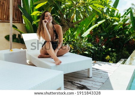 The smiling girl bites the finger of her thumb and sits on the standing on the terrace against a backdrop of tropical plants. High quality photo