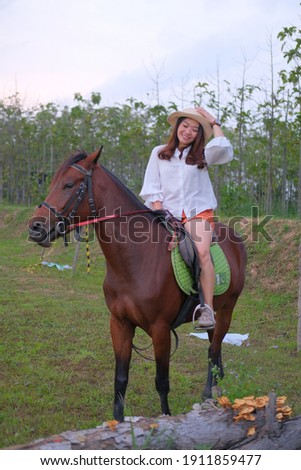 concept beauty photography and animal photography, young and beauty asian lady or woman on white shirt and orange pants with brown horse with blurry trees background.