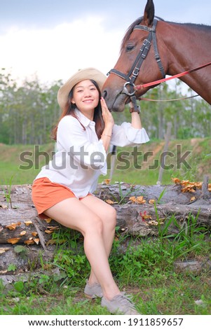concept beauty photography and animal photography, young and beauty asian lady or woman on white shirt and orange pants with brown horse with blurry trees background.