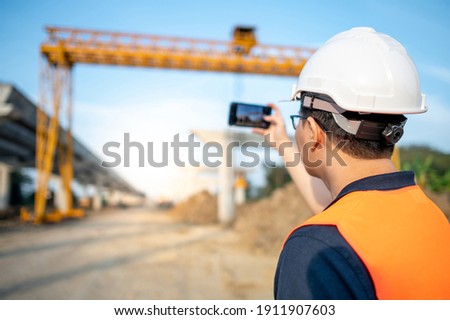 Smart Asian worker man or male civil engineer with protective safety helmet and reflective vest using using smartphone for taking photo at construction site.