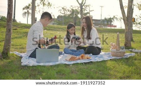 Happy family asian and father is playing Ukulele while little take a picture enjoyed ourselves together during picnicking on a picnic cloth in the green garden. Family enjoying sunny fall day.