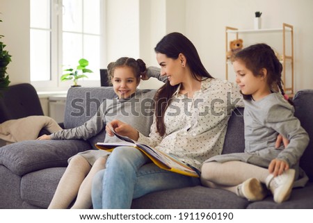 Young mother with her little twin daughters are reading fairy tales and looking at pictures in a book while sitting on a sofa at home. Young family have fun together during the weekend.