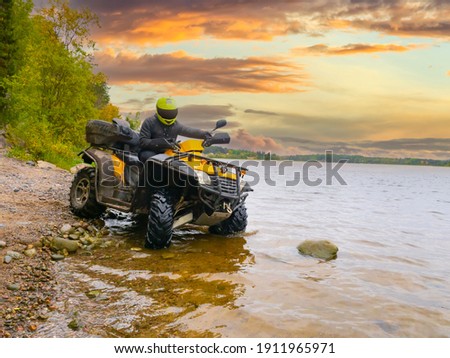 Quad bike on the river bank. The ATV driver drove into water. Quad bike on the background sunset. A man travels on a quad bike. A yellow ATV stands in lake. Concept - outdoor activity.