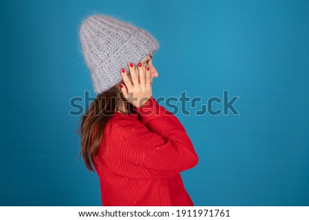 Young woman in warm knitted hat shows her red nails on the blue background, side view. Winter cozy clothes concept. High quality photo
