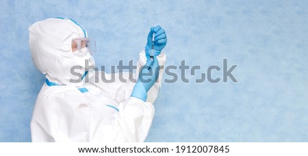 
Medical worker in a white protective suit on a blue background. Wear a respirator and gloves. Prepares a syringe for a vaccine set. Place for an inscription. Panorama.