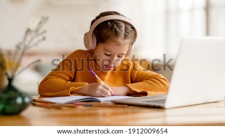 Focused serious elementary  little girl in headphones   making notes in notepad while communicating with teacher through video chat app during online education at home Royalty-Free Stock Photo #1912009654