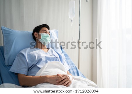 Asian male patient lying on bed with face mask in recovery room in hospital ward. All people wearing mask to prevent covid19 virus infection during coronavirus pandemic. The man feels lonely and bore. Royalty-Free Stock Photo #1912026094