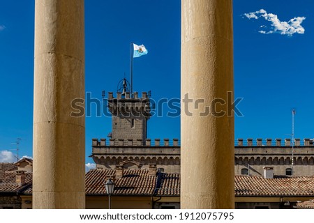 The flag of the Republic of San Marino flutters in the blue sky over the Palazzo Pubblico, framed by the columns of the basilica
