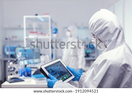 Concentrated medical researcher using digital tablet dressed in protective suit against infection with coronavirus. Team of scientists conducting vaccine development using high tech technology for