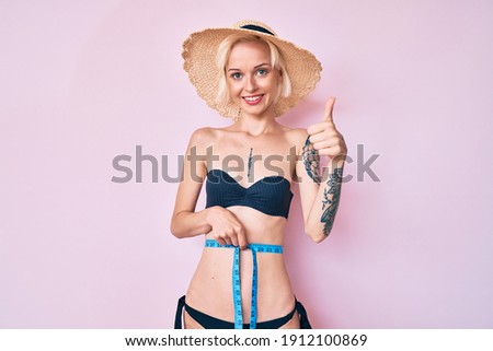 Young blonde woman with tattoo wearing bikini using tape measure smiling happy and positive, thumb up doing excellent and approval sign 