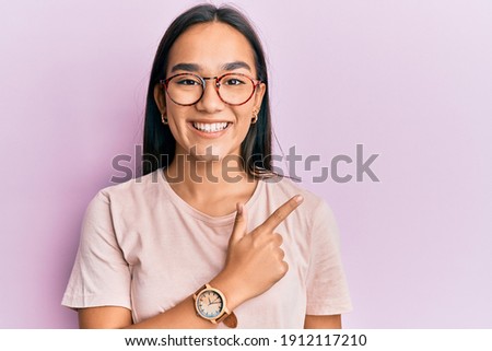 Young asian woman wearing casual clothes and glasses smiling cheerful pointing with hand and finger up to the side 