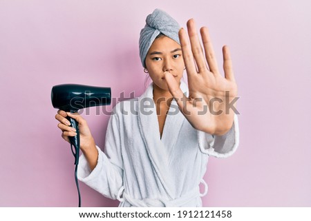 Young chinese woman wearing shower towel cap and bathrobe holding dryer with open hand doing stop sign with serious and confident expression, defense gesture 