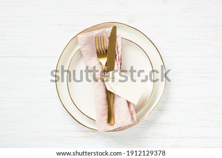 Golden rim plates and golden cutlery on pink napkin with tag on white table. Top view.