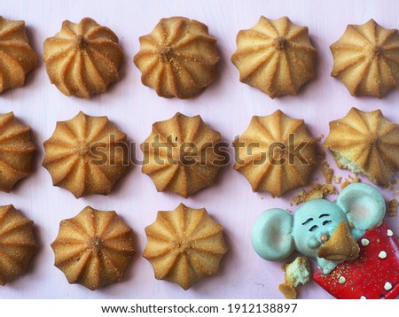 Small round cookies lie in rows on the pink surface, in the lower right corner instead of cookies there is a toy mouse and cookie crumbs 