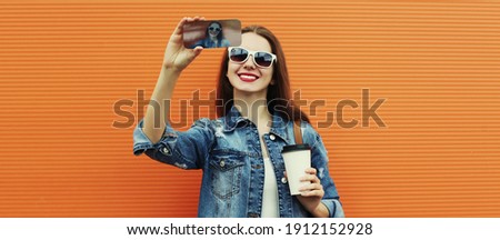 Portrait of smiling woman taking a selfie picture by smartphone wearing a denim jacket on a orange background