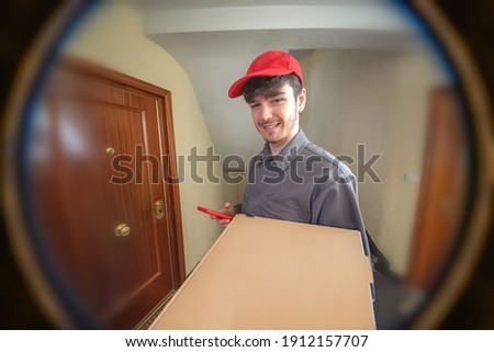 Fast food delivery man with pizza box in his hands delivering the order, dressed in uniform and smiling, customer looking through the peephole Royalty-Free Stock Photo #1912157707