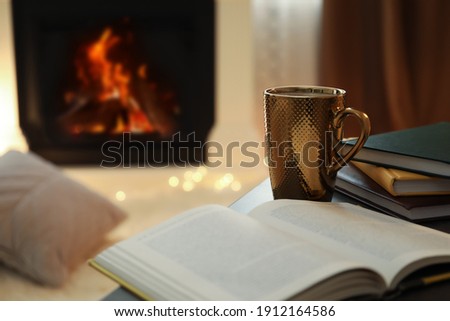 Cup of hot drink and book on table near fireplace at home. Cozy atmosphere