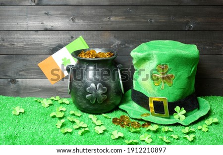 Cast iron pot with treasure, irish flag with clover leaf and leprehaun hat, clover leaves and coins on green grass, dark wooden planks background. Saint Patricks Day banner, poster, flyer, invitation
