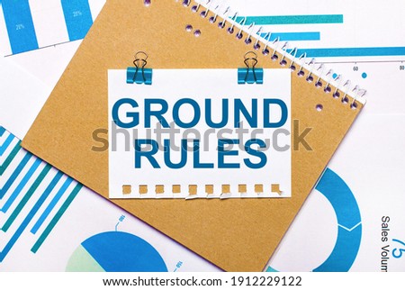 On the desktop are blue and light blue graphs and diagrams, a brown notebook and a sheet of paper with blue clips and GROUND RULES text. View from above. Business concept