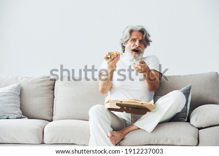 Soccer fan with pizza watches match. Senior stylish modern man with grey hair and beard indoors.