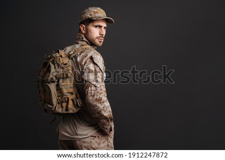 Confident masculine military man in uniform posing with backpack isolated over black background