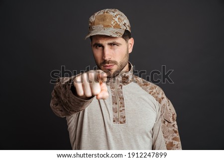 Confident masculine military man looking and pointing finger at camera isolated over black background