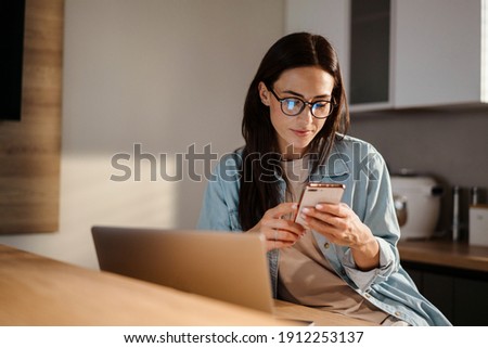 Serious charming woman using smartphone while working with laptop at home Royalty-Free Stock Photo #1912253137