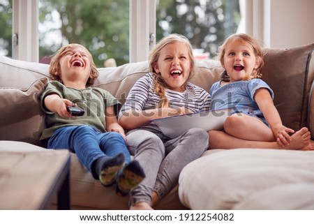 Three Children Sitting On Sofa At Home Laughing And Watching TV With Popcorn Royalty-Free Stock Photo #1912254082