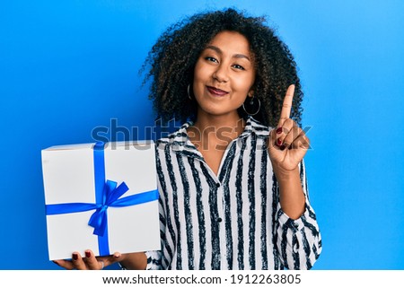 Beautiful african american woman with afro hair holding gift smiling with an idea or question pointing finger with happy face, number one 