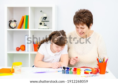 A girl with down syndrome is studying at home with her mother. Girl and mother sitting at the table draw a picture at home.