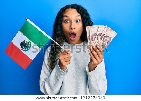 Young latin woman holding mexico flag and mexican pesos banknotes afraid and shocked with surprise and amazed expression, fear and excited face. 